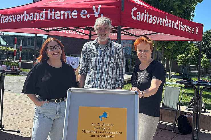Drei Personen stehen vor einem Caritas-Pavillon.