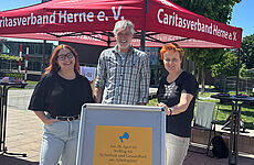 Drei Personen stehen vor einem Caritas-Pavillon.