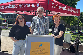 Drei Personen stehen vor einem Caritas-Pavillon.