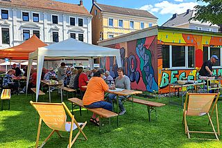 Menschen sitzen auf Bänken vor einem bunt bemalten Container.