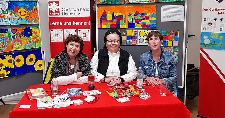 Drei Frauen sitzen vor einer Stellwand mit bunten Bildern.