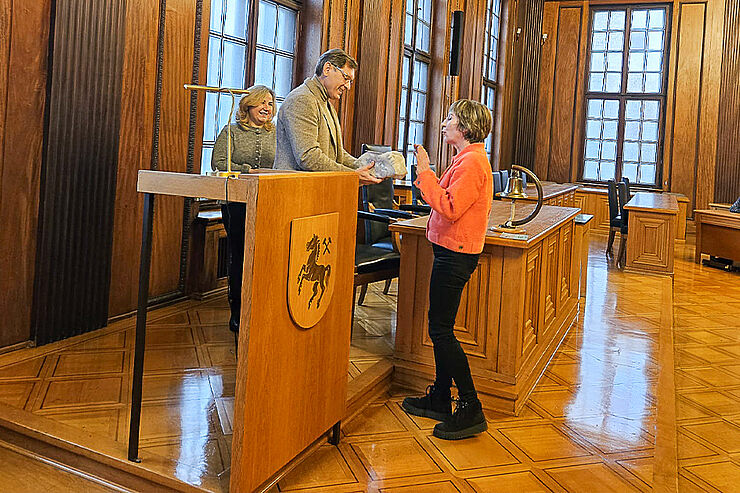 Eine Frau übergibt einem Mann ein Geschenk, im Hintergrund steht eine weitere Frau.