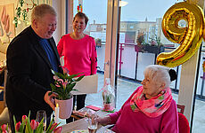 Ein Mann überreicht einer älteren Dame Blumen, im Hintergrund lächelt eine Frau.