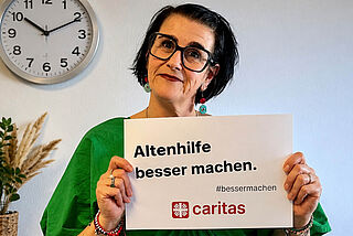 Eine Frau hält ein Schild mit der Aufschrift "Altenhilfe besser machen" in die Kamera.