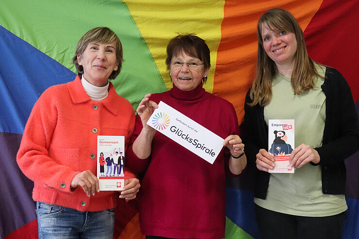 Drei Frauen stehen nebeneinander vor einer Regenbogenflagge.