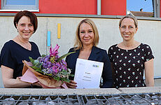 Drei Frauen stehen mit einem Blumenstrauß und einem Dokument in der Hand zusammen.