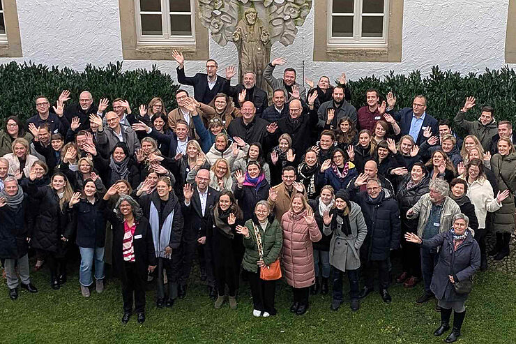 Eine große Gruppe Menschen steht auf einer Wiese zusammen und winkt.