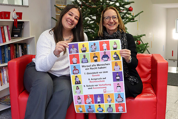 Zwei Frauen sitzen nebeneinander und halten ein buntes Plakat.