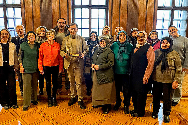 Eine große Gruppe Menschen steht vor großen Glasfenstern, in der Mitte steht ein Mann mit einem Stiftehalter in der Hand.
