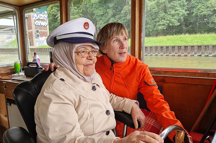 Eine Frau mit Kapitänsmütze sitzt an einem Steuerrad, eine Frau steht daneben.