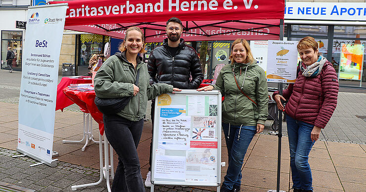 Vier Menschen stehen vor einem Pavillon um einen Aufsteller herum.