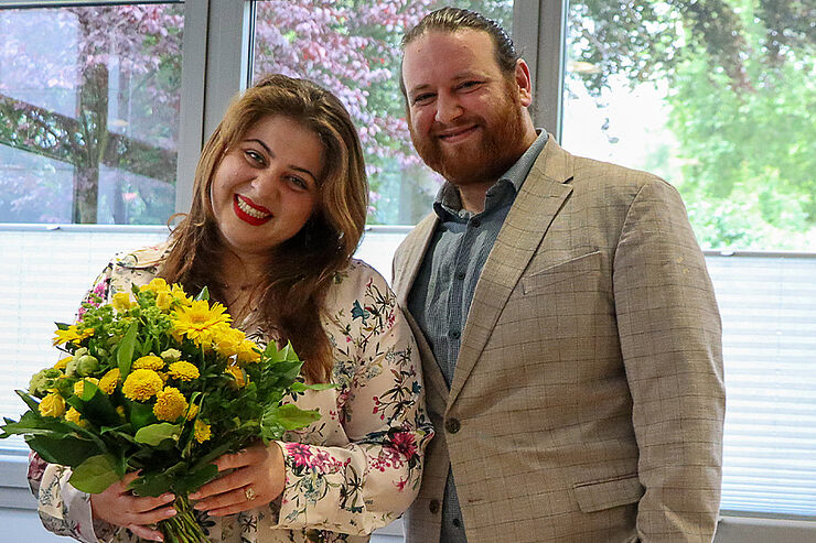 Eine Frau mit Blumen und ein Mann stehen nebeneinander und lächeln.