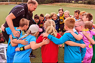 Ein Mann und eine Gruppe Kinder stehen Arm in Arm im Kreis und lachen.