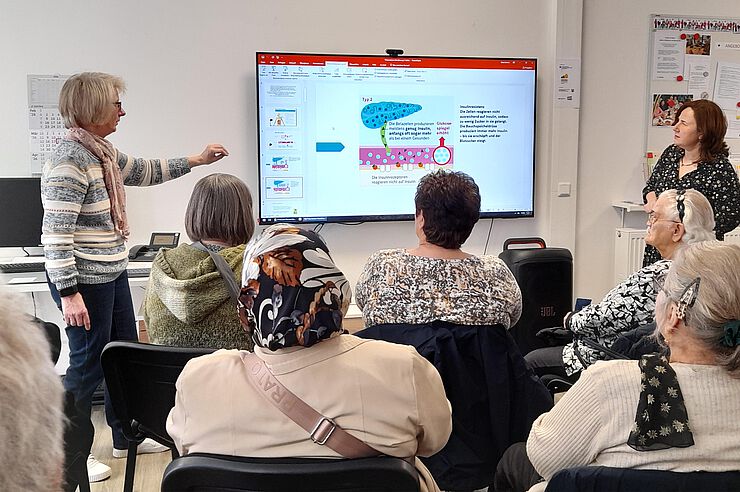 Zwei Frauen stehen neben einem Bildschirm, mehrere Menschen sitzen davor.