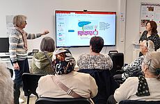 Zwei Frauen stehen neben einem Bildschirm, mehrere Menschen sitzen davor.