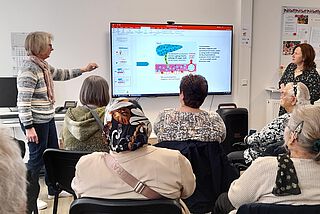 Zwei Frauen stehen neben einem Bildschirm, mehrere Menschen sitzen davor.